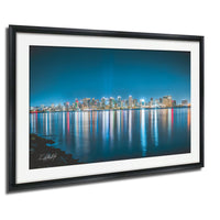 A framed photograph depicting a city skyline at night, with the buildings' lights reflected in the water below.