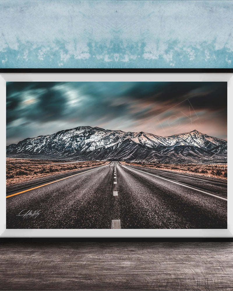 A framed photograph depicting a long, straight road leading towards a snow-capped mountain range under a dramatic, cloudy sky.