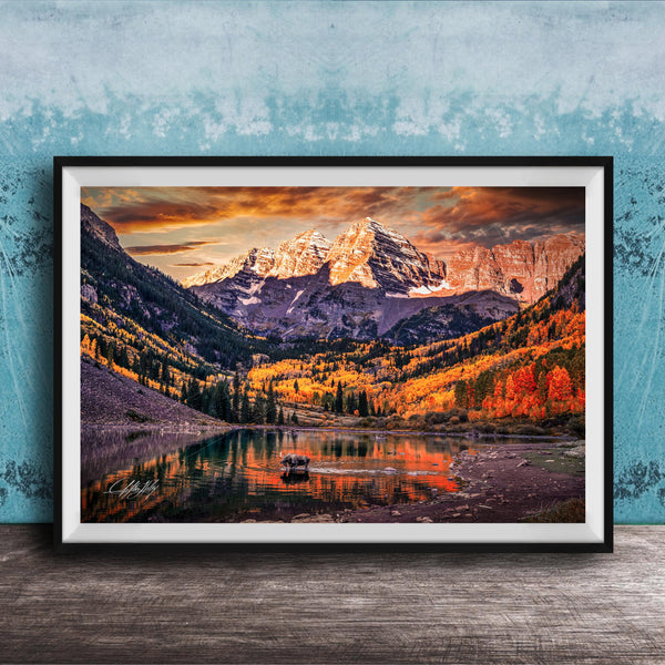 a breathtaking mountain landscape with a lake in the foreground, surrounded by vibrant autumn foliage. The mountains are covered in snow, creating a stunning contrast against the colorful sky. The scene is framed in a black frame and displayed on a wooden surface, adding to the rustic charm of the setting.