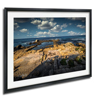 The image is a framed photograph of a rocky coastal landscape with a clear blue sky and white clouds.