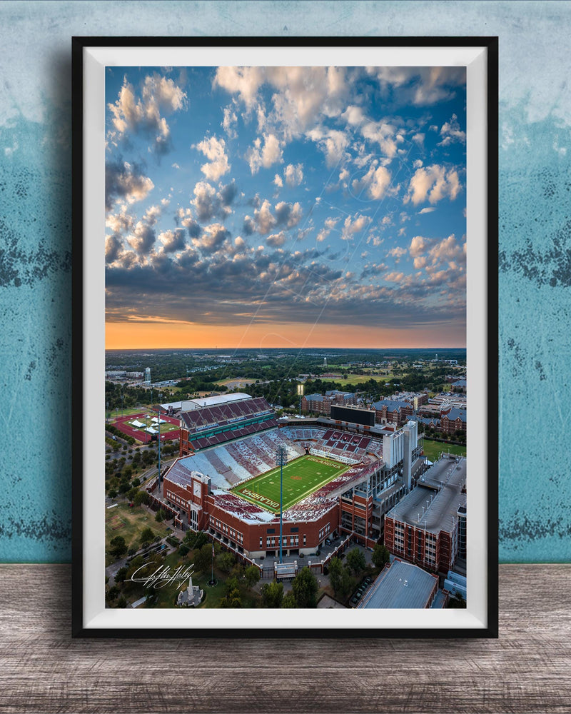 The image is an aerial view of a large sports stadium with a football field, surrounded by buildings and a cloudy sky.