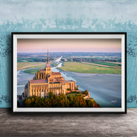 a large, ornate cathedral with a tall spire, situated on a cliff overlooking a body of water. The cathedral is surrounded by lush greenery, creating a picturesque scene. The photograph is framed and displayed on a wooden surface, with a blue wall in the background.