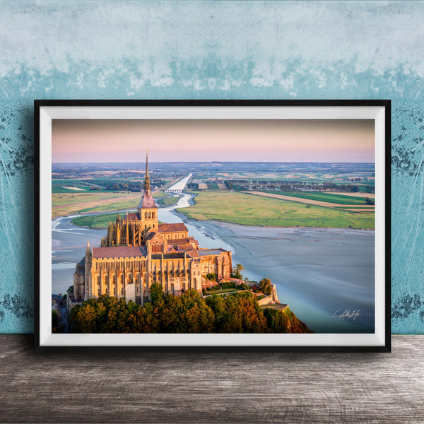 a large, ornate cathedral with a tall spire, situated on a cliff overlooking a body of water. The cathedral is surrounded by lush greenery, creating a picturesque scene. The photograph is framed and displayed on a wooden surface, with a blue wall in the background.