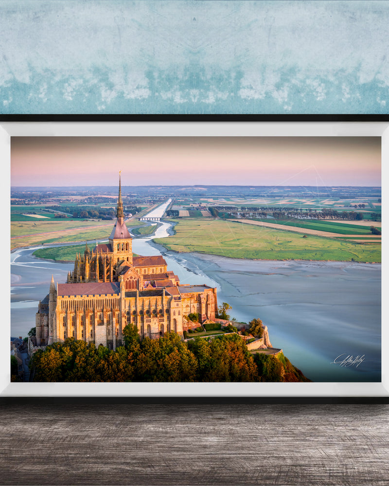 a large, ornate cathedral with a tall spire, situated on a cliff overlooking a body of water. The cathedral is surrounded by lush greenery, creating a picturesque scene. The photograph is framed and displayed on a wooden surface, with a blue wall in the background.