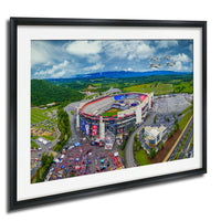 The image is an aerial view of a large stadium surrounded by a large field, with a few buildings and vehicles visible in the vicinity.