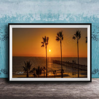 A framed photograph of a serene beach scene at sunset, with palm trees silhouetted against the warm orange sky.