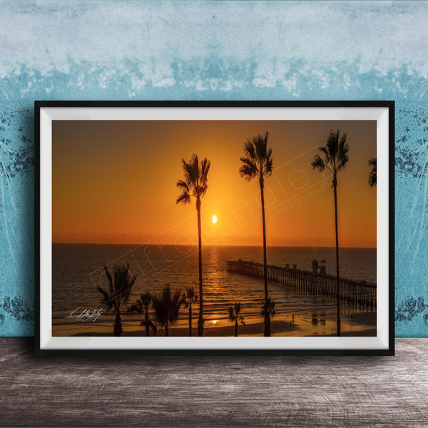 A framed photograph of a serene beach scene at sunset, with palm trees silhouetted against the warm orange sky.
