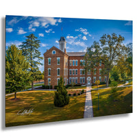 A large brick building with a clock tower, surrounded by trees and a grassy lawn, set against a blue sky with scattered clouds.