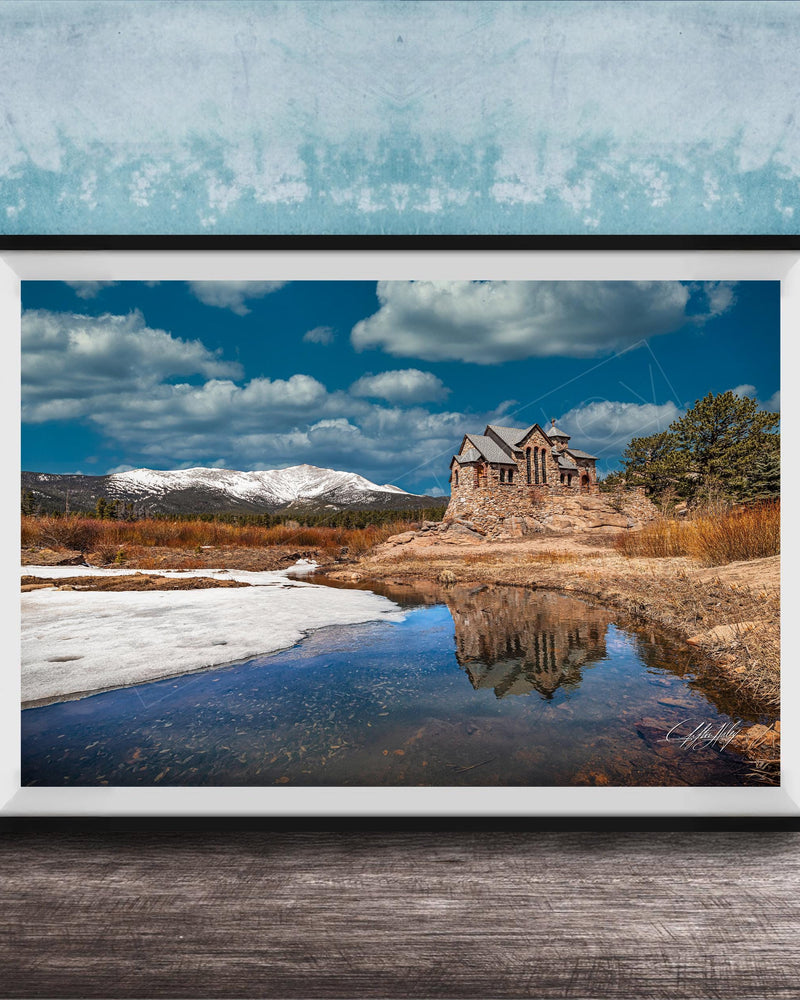 A framed photograph depicting a stone building reflected in a calm body of water, with snow-capped mountains in the background and a cloudy blue sky above.