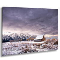 A snowy field with a wooden barn in the foreground, set against a backdrop of snow-capped mountains under a dramatic cloudy sky.