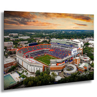 An aerial view of a large sports stadium with a crowd of spectators, surrounded by a cityscape and a colorful sunset sky.