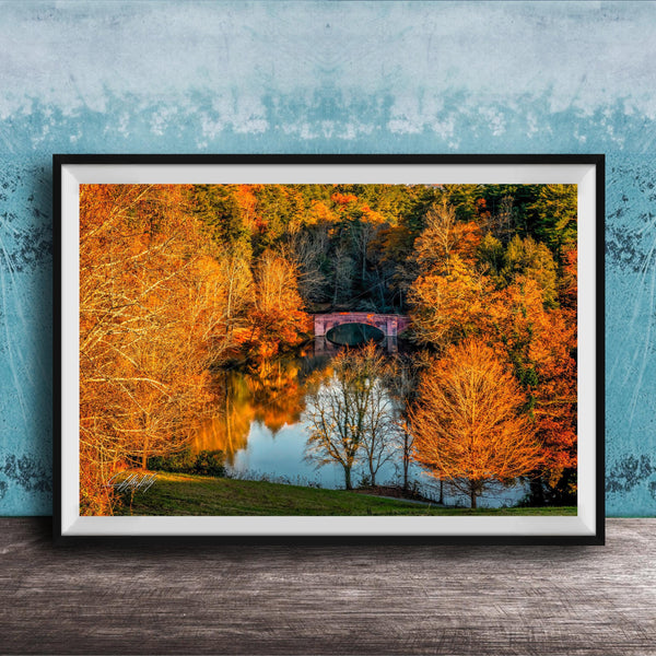 A framed photograph depicting a scenic autumn landscape with a red bridge spanning a tranquil lake surrounded by vibrant orange and yellow foliage.