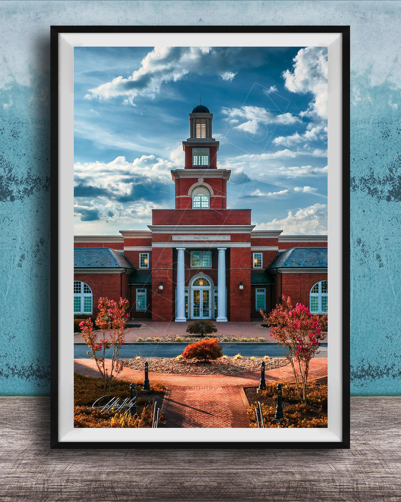 A framed photograph of a red brick building with a prominent clock tower, set against a blue sky with clouds.