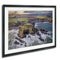 The image is a framed aerial view of a coastal landscape, featuring a rocky outcrop, a small village or settlement, and the ocean with crashing waves.