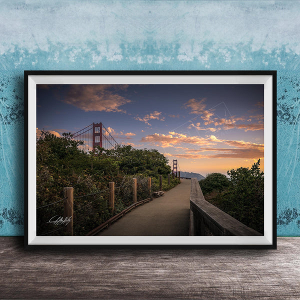 A framed photograph depicting a scenic view of the Golden Gate Bridge at sunset, with a wooden walkway leading towards the bridge and a colorful sky in the background.