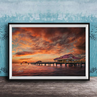 A framed photograph depicting a serene sunset over a wooden pier, with a small building on the pier and a calm body of water in the foreground.