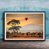 A framed photograph depicting a serene landscape with a lone tree, a herd of elephants, and a red hot air balloon floating in the sky, all set against a backdrop of a majestic mountain range.