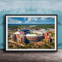 The image is a framed aerial view of a large sports stadium, showcasing its distinctive architecture and surrounding landscape.