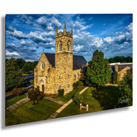 A stone church with a tall tower, surrounded by trees and a grassy lawn, set against a blue sky with clouds.