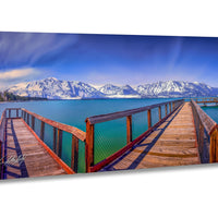 A wooden pier extends into a serene lake, with snow-capped mountains rising in the distance against a clear blue sky.