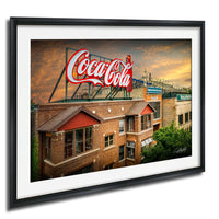 A framed image of a large Coca-Cola sign on top of a brick building, with a cloudy sky in the background.
