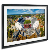 The image is an aerial view of a large sports stadium with a football field, surrounded by buildings and greenery.