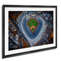 An aerial view of a baseball stadium with a green field and red diamond, surrounded by a cityscape with buildings and trees.