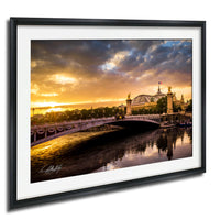 a picturesque scene of a bridge spanning a river, with a large domed building on the other side. The sky is filled with clouds, and the sun is setting, casting a warm glow over the entire scene. The bridge is adorned with statues, adding to its majestic appearance. The image is framed and appears to be a painting or photograph, capturing the beauty of the landmark.