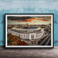 The image is a framed photograph of the New York Yankees stadium, which is a large, circular stadium located in the city of New York. The photograph captures the stadium's distinctive architecture and surrounding cityscape, with a dramatic sky in the background.