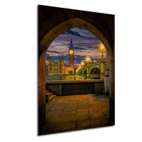 a picturesque view of the Palace of Westminster and the Elizabeth Tower, also known as Big Ben, in London, England, with the River Thames in the foreground. The scene is captured through an archway, creating a sense of depth and perspective. The sky is illuminated by the warm glow of the setting sun, casting a beautiful light over the iconic landmarks.