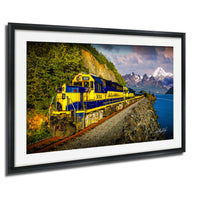 A yellow and blue train engine, numbered 3001, is traveling along a track that runs along the edge of a cliff overlooking a body of water and snow-capped mountains in the distance.
