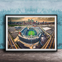 An aerial view of a large sports stadium surrounded by a city skyline, with the stadium's green field and surrounding parking lots visible.