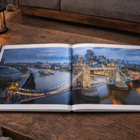 An open book displaying a scenic view of London, with the iconic Tower Bridge and other cityscape visible. The book is placed on a wooden surface, with a lit candle in the background and some fallen leaves nearby.