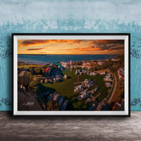 The image is a framed aerial view of a harbor with boats docked, set against a backdrop of a colorful sunset sky.