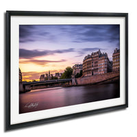 a picturesque cityscape with a river running through it, featuring a bridge and buildings along the riverbanks. The sky is painted in warm hues of orange and pink, creating a serene and tranquil atmosphere. The image is framed and appears to be a high-quality, professional-looking photograph.
