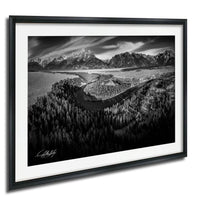 A black and white landscape photograph depicting a winding road surrounded by a forested area, with snow-capped mountains in the background.
