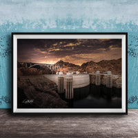 A framed photograph depicting a large dam with a bridge spanning across it, set against a dramatic sky with clouds and a sunset.