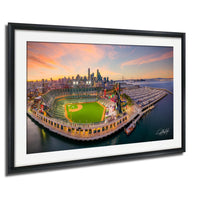 The image is a framed aerial view of a baseball stadium with a city skyline in the background, set against a colorful sunset sky.