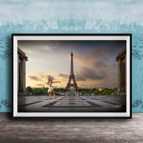 A framed photograph of a woman in a white dress dancing in front of the Eiffel Tower in Paris, France, with a blue sky and clouds in the background.
