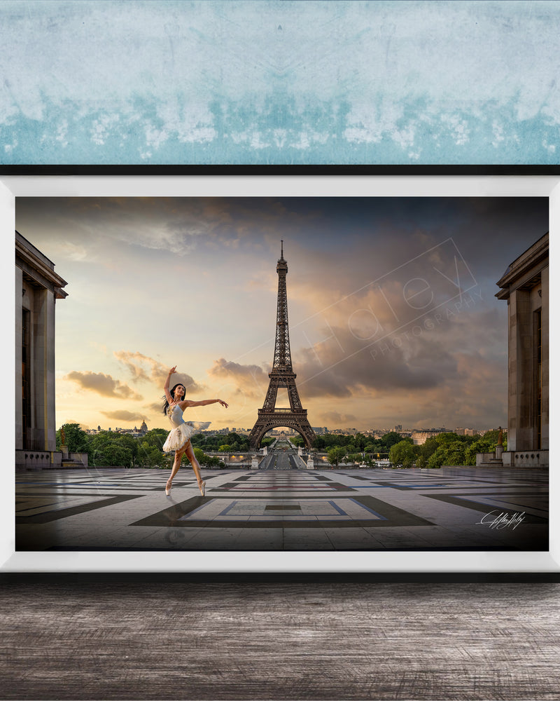 A framed photograph of a woman in a white dress dancing in front of the Eiffel Tower in Paris, France, with a blue sky and clouds in the background.