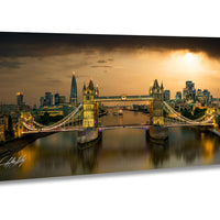 A panoramic view of the London skyline at night, featuring the iconic Tower Bridge spanning the River Thames. The bridge is illuminated with lights, creating a striking contrast against the dark sky. The cityscape is reflected in the calm waters of the river, adding to the overall beauty of the scene.