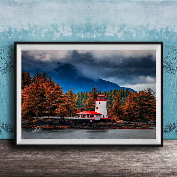 A framed photograph depicting a lighthouse surrounded by a forest with autumn-colored trees, set against a cloudy sky.