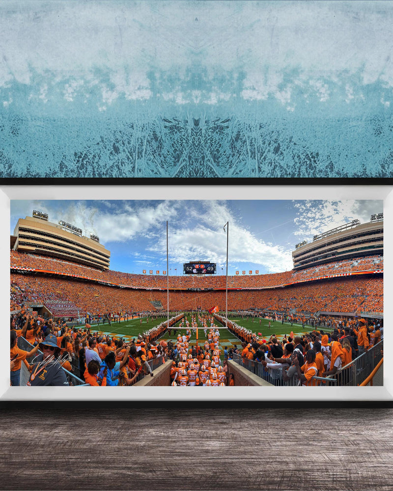 A framed photograph of a crowded football stadium with a large crowd of spectators, a large scoreboard, and a large field.