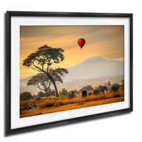 A framed photograph depicting a serene landscape with a lone tree, a herd of elephants, and a hot air balloon in the sky, all set against a warm, golden sunset.