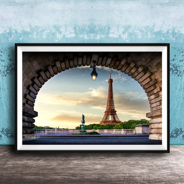 A framed photograph of the Eiffel Tower in Paris, France, with the tower visible through an archway. The sky is blue with clouds, and the sun is setting, creating a picturesque scene.