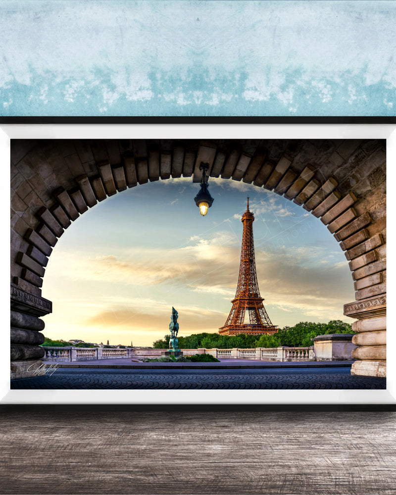 A framed photograph of the Eiffel Tower in Paris, France, with the tower visible through an archway. The sky is blue with clouds, and the sun is setting, creating a picturesque scene.