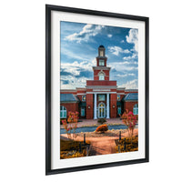 A framed photograph of a red brick building with a tower, surrounded by a well-maintained garden with trees and flowers.