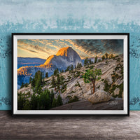 The image is a framed photograph depicting a mountainous landscape with a large mountain peak in the background, surrounded by trees and rocky terrain. The sky is filled with warm, golden light, creating a picturesque and serene atmosphere.