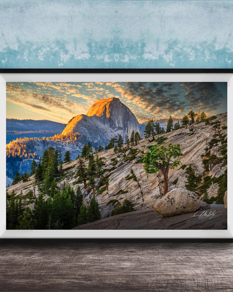 The image is a framed photograph depicting a mountainous landscape with a large mountain peak in the background, surrounded by trees and rocky terrain. The sky is filled with warm, golden light, creating a picturesque and serene atmosphere.