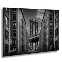 A black and white photograph of the Manhattan Bridge, a suspension bridge with two towers and a walkway, spanning between two tall buildings in an urban setting.
